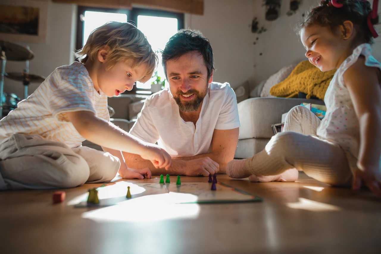 © KWB, Halfpoint Ein Vater spielt mit seinen zwei Kindern ein Brettspiel auf dem Wohnzimmerboden.