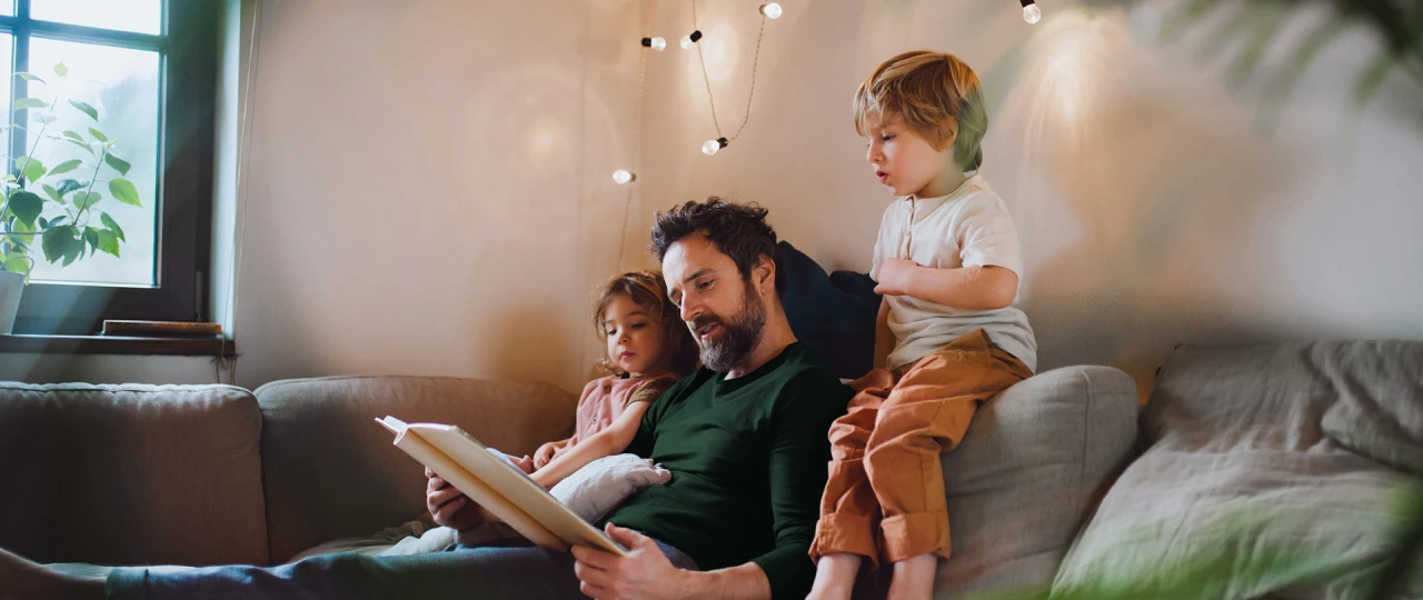 © KWB, Halfpoint Ein Vater sitzt mit seinen zwei Kindern gemütlich auf dem Sofa und liest eine Geschichte vor.