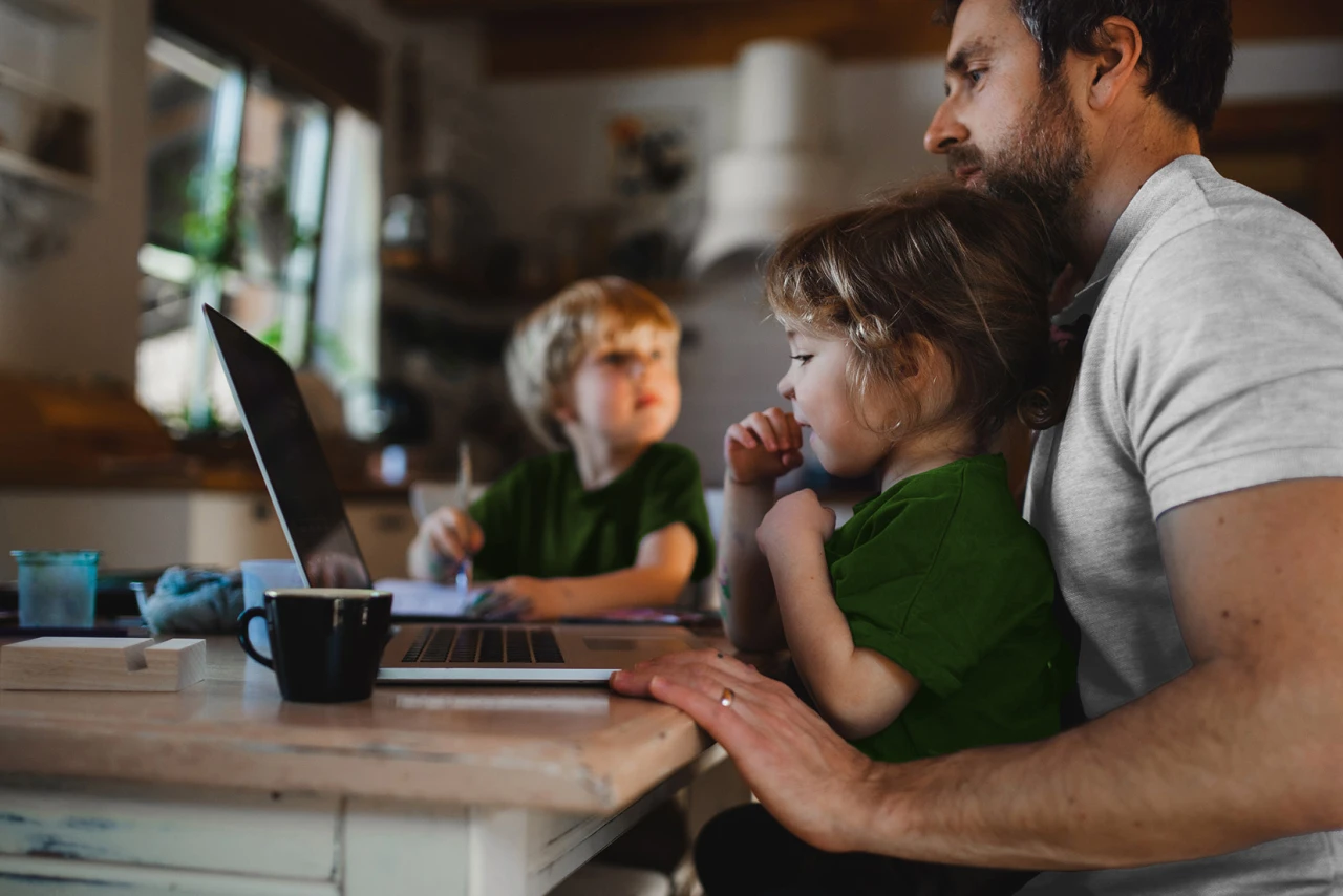 © KWB, Halfpoint Vater sitzt mit zwei Kindern am Küchentisch vor einem Laptop, während sie gemeinsam konzentriert arbeiten.