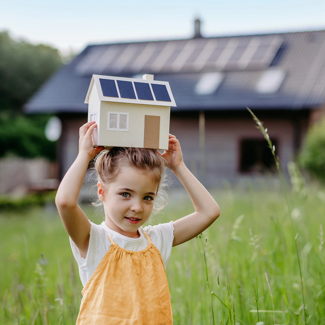 © KWB, Halfpoint Junges Maedchen haelt Kartonhaus mit PV-Modulen über dem Kopf und laechelt.
