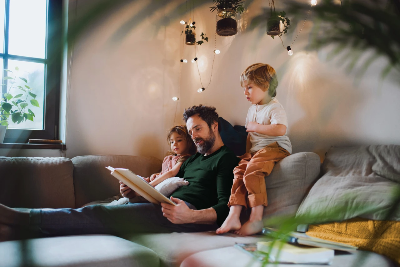 © KWB, Halfpoint Ein Vater sitzt mit seinen zwei Kindern gemütlich auf dem Sofa und liest eine Geschichte vor.