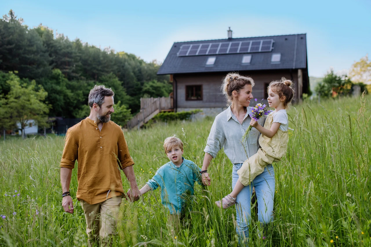© KWB, Halfpoint Eine Familie geht gemeinsam durch eine blühende Wiese vor ihrem modernen Holzhaus mit Photovoltaikanlage am Dach.