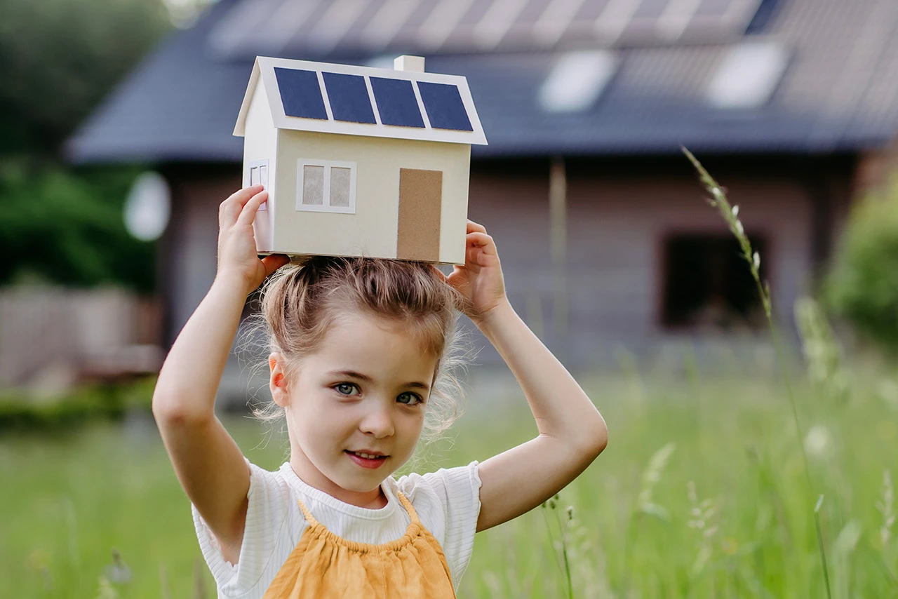 © KWB, Halfpoint Junges Maedchen haelt Kartonhaus mit PV-Modulen über dem Kopf und laechelt.