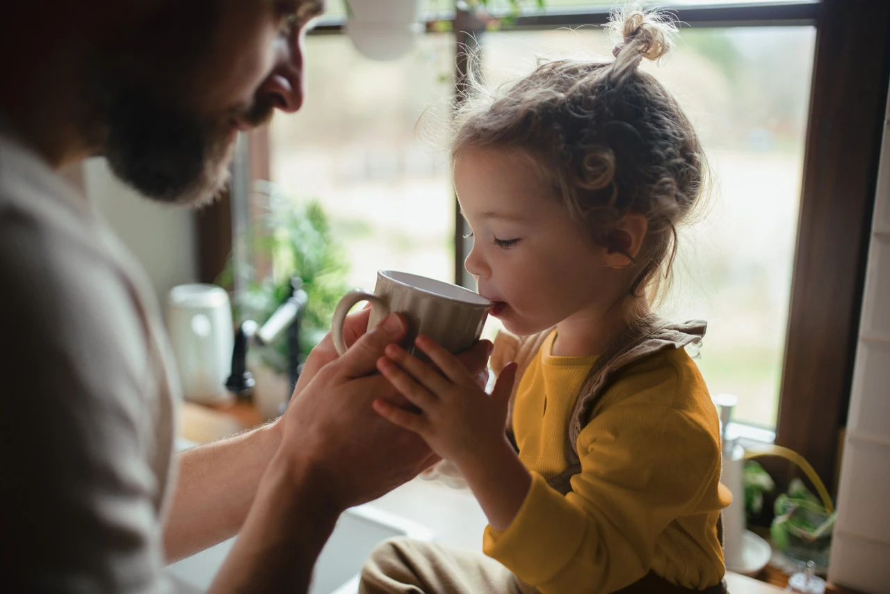 © KWB, Halfpoint Ein kleines Kind trinkt aus einer Tasse, die vom Vater gehalten wird.