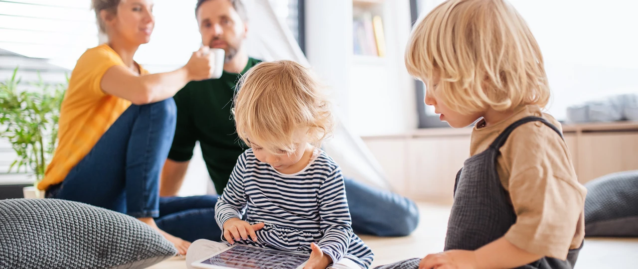 © KWB, Halfpoint Zwei kleine Kinder spielen auf dem Boden im Wohnzimmer, eines mit einem Tablet. Im Hintergrund sitzen die Eltern entspannt beisammen und trinken Kaffee.