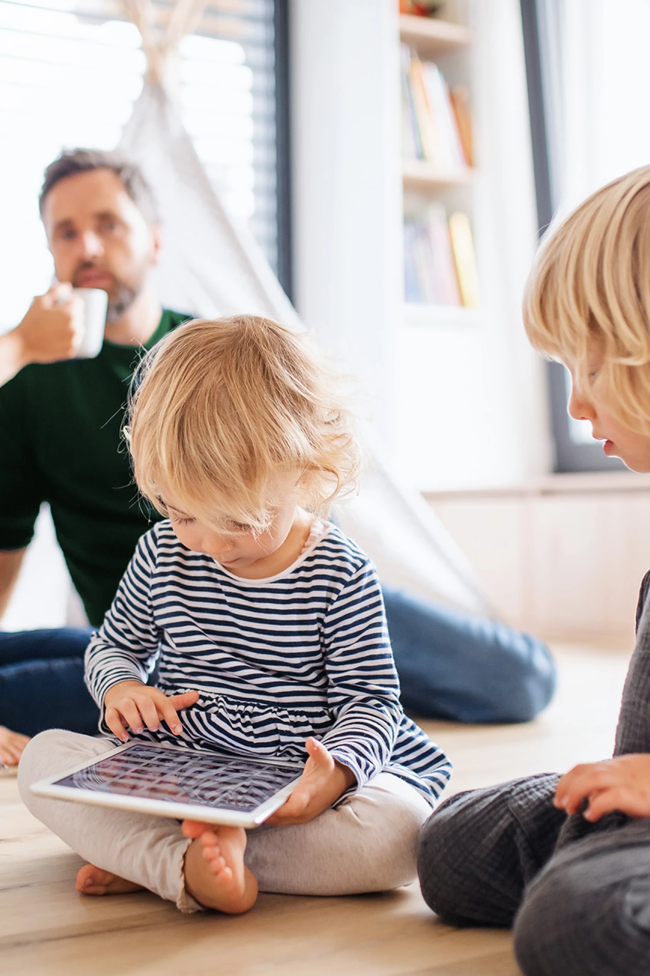 © KWB, Halfpoint Zwei kleine Kinder spielen auf dem Boden im Wohnzimmer, eines mit einem Tablet. Im Hintergrund sitzen die Eltern entspannt beisammen und trinken Kaffee.